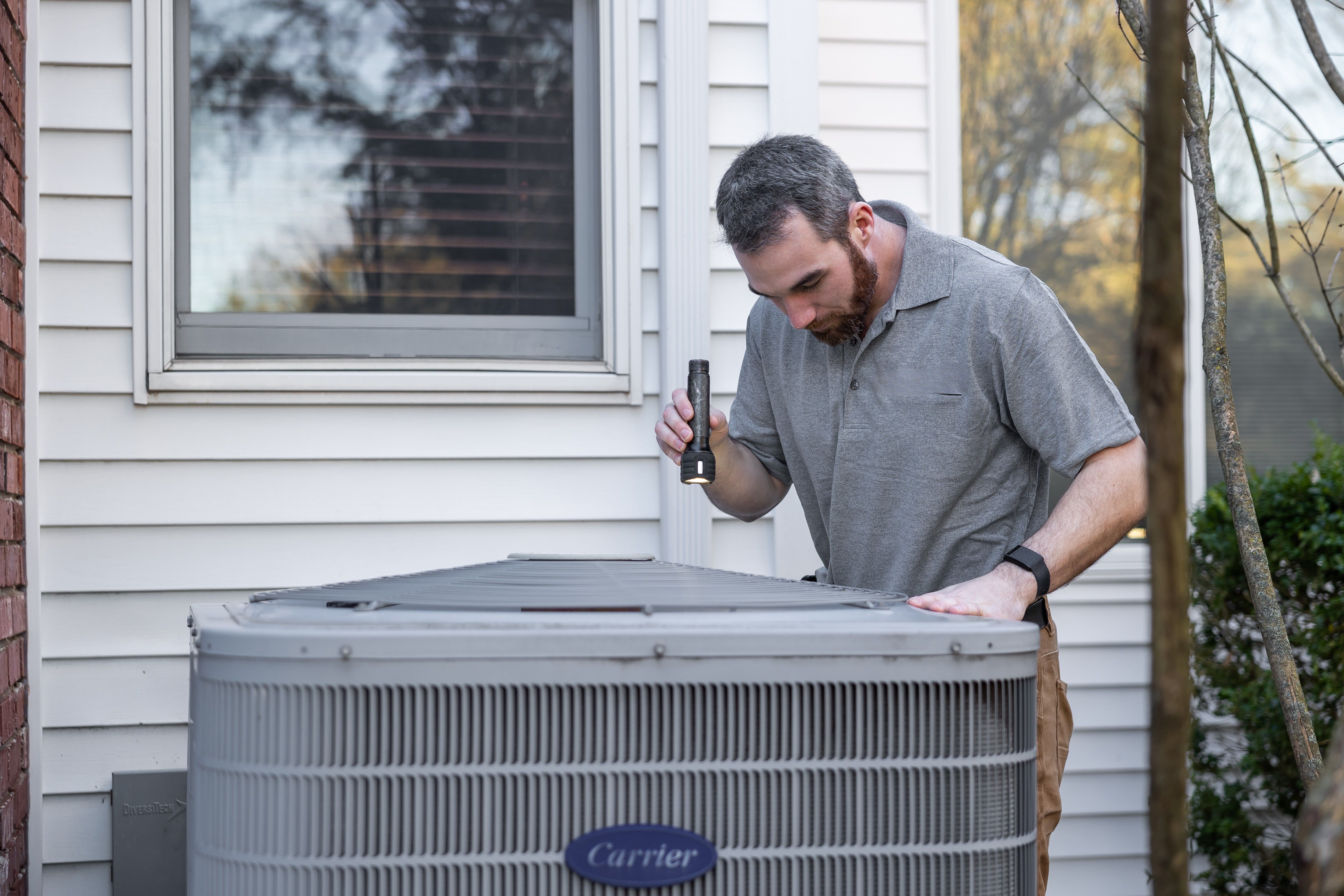 Technician inspecting an outdoor HVAC condenser beside a house