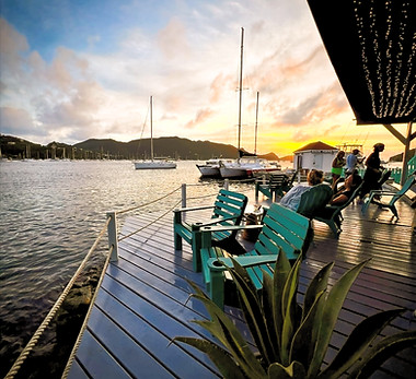Tranquil waterfront at sunset with teal Adirondack chairs on a wooden deck, people relaxing, sailboats on the calm water, and a backdrop of mountains. At Bequia Marina Cocktail Lab Bar.