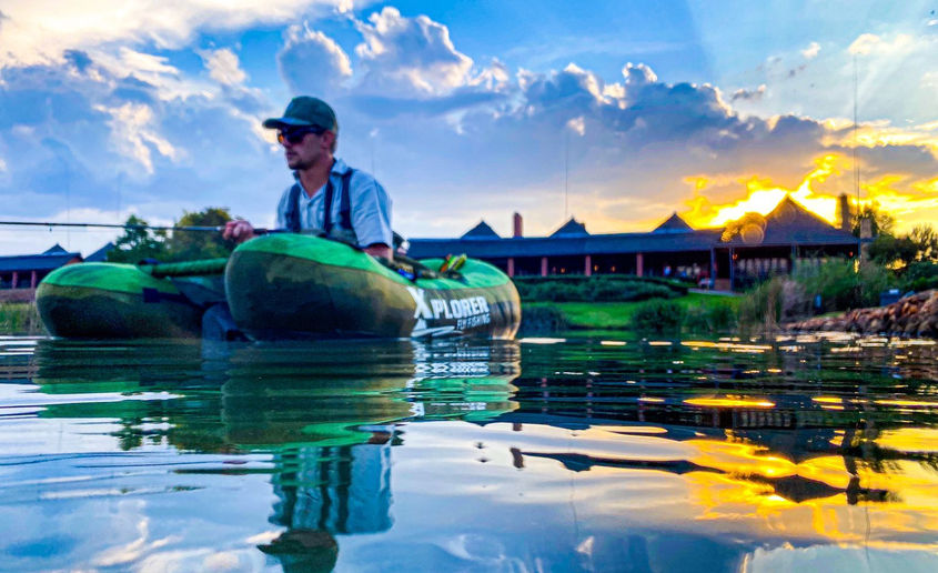 A man fly-fishing while sitting in a boat on a lake that reflects the beautiful sunrise sky.