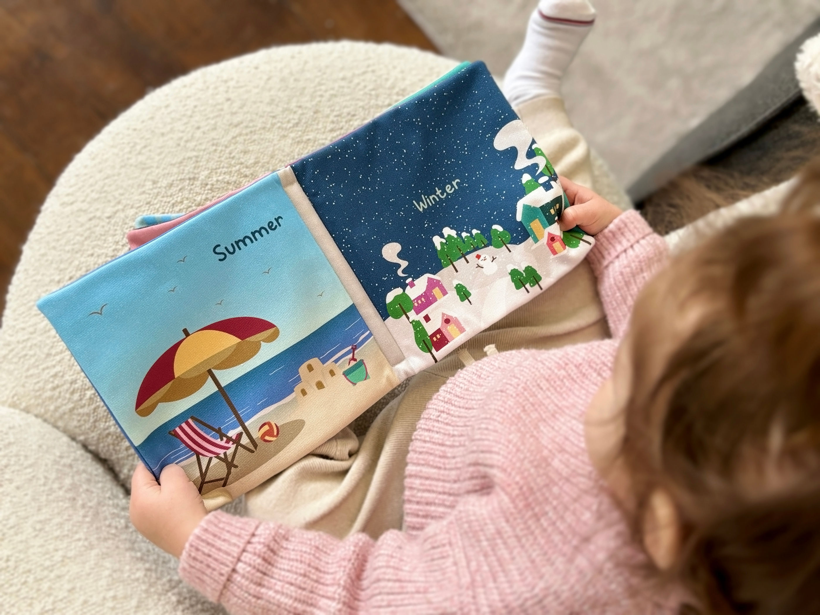 Little girl holding My First Book Of Opposites and showing pages with summer and winter illustrations