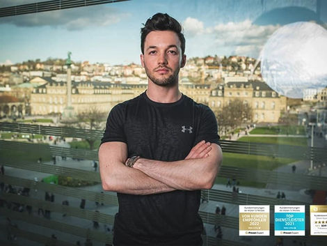 Dimitri Rutansky in schwarzem T-Shirt steht mit verschränkten Armen vor einem Fenster. Im Hintergrund ist eine Stadtlandschaft zu sehen.
