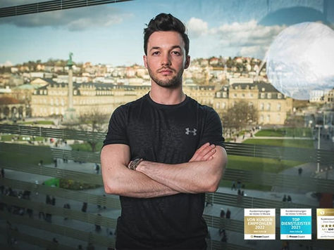 Dimitri Rutansky in schwarzem T-Shirt steht mit verschränkten Armen vor einem Fenster. Im Hintergrund ist eine Stadtlandschaft zu sehen.