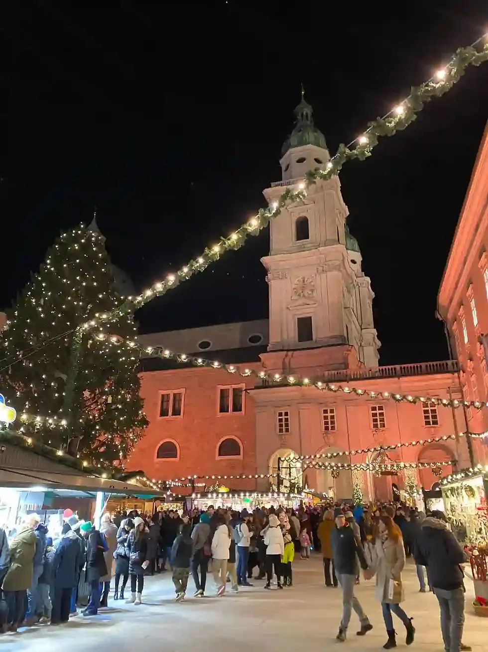 Crowded Christmas market at night in front of Salzburg Cathedral, with a lit Christmas tree and garlands of lights