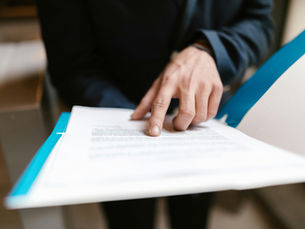 A person in a dark suit points at text on a document with a blue folder. The setting is indoors, creating a focused and professional mood.