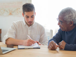Young man writing on clipboard discusses with older man at wooden table, laptop open. Bright setting, focused mood.
