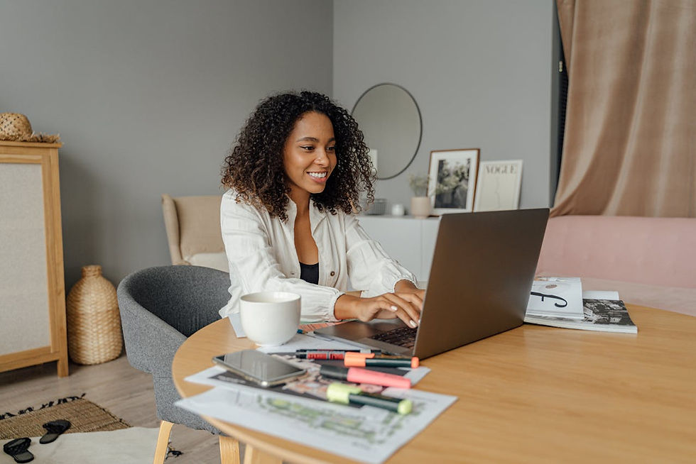 Smiling woman using a laptop at a wooden table with papers, pens, and a mug. Softly lit room with framed art and a round mirror on a shelf.