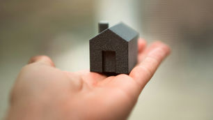 Hand holding a small, black house model against a blurred background. The model has simple geometric lines, evoking a minimalist mood.