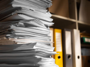 Stack of papers on a desk with yellow binders in the background, evoking a busy office environment. Soft lighting creates contrast.