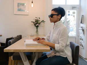 A woman in sunglasses reads a Braille book at a table in a bright room with flowers, a hanging light, and framed art. Casual and serene mood.