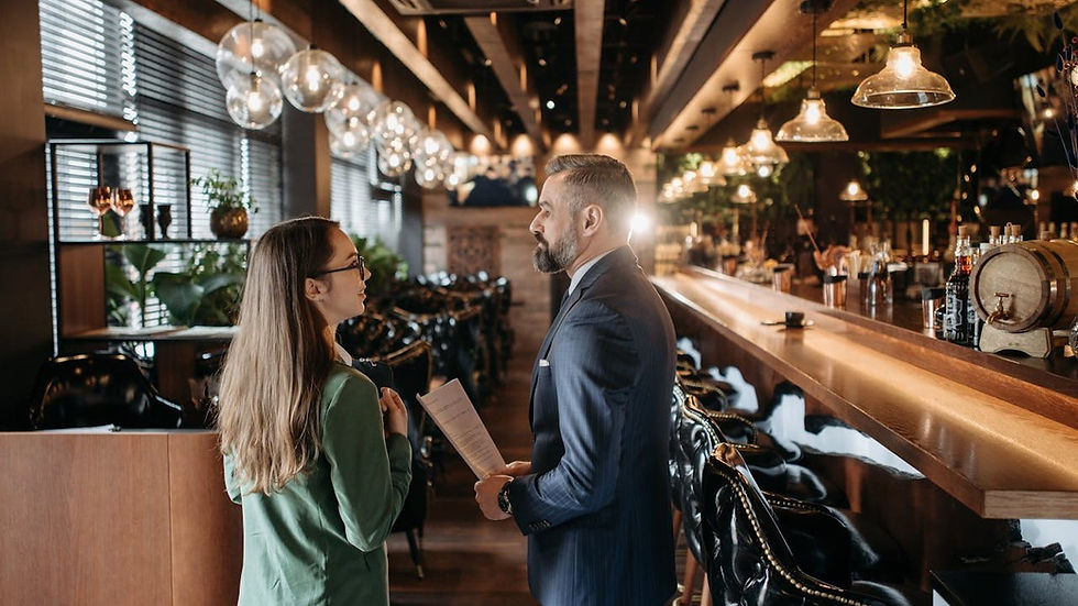 Two colleagues talk in a stylish, warmly lit bar; the man holds a menu beside the long wooden counter.