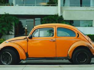 Orange vintage car parked on street with lush greenery and modern building in background. Car shows signs of wear, creating a nostalgic mood.