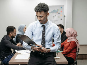 Man in shirt and tie holds clipboard in office meeting while colleagues sit around table, focused and serious.