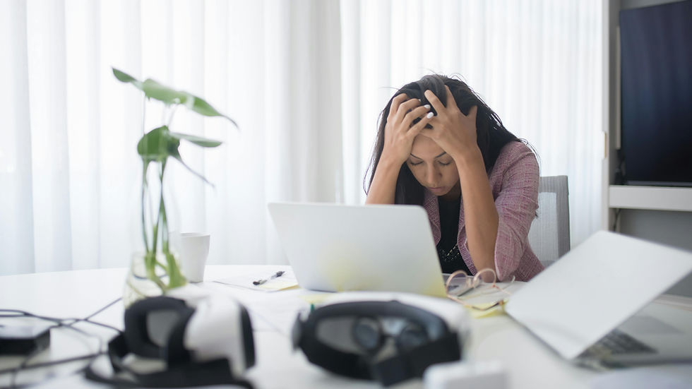 Woman sits at a desk, head in hands, stressed, overwhelmed a. Laptop open, VR headsets, and plant visible. Bright curtains in the background.