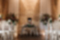two pedestals and an arrangement on a small table of bright flowers against a yellow cotswold stone wall with chairs either side of aisle.