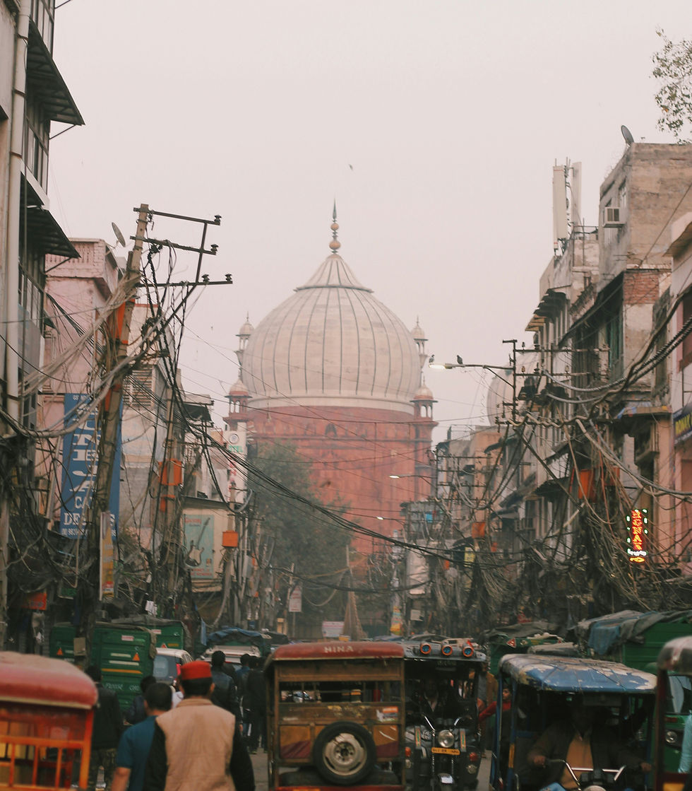 Busy street scene with people, rickshaws, and tangled wires. Large domed building in the background. Overcast sky and bustling atmosphere.