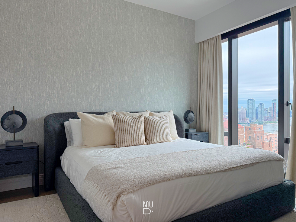 Luxury primary bedroom with custom upholstered bed, neutral bedding, and skyline views through floor-to-ceiling windows in a modern New York residence.