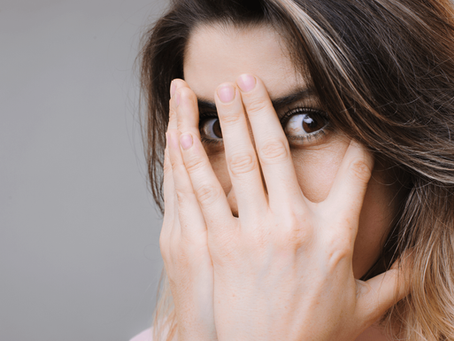 Eine Frau hält die Hände vor ihr Gesicht vor Scham. Sie sieht direkt in die Kamera durch ihre Finger. 