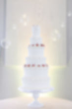 A white five-tiered cake with ornate icing and strawberries on a stand. Soft lighting and hanging decorations create an elegant ambiance. Venue is Rancho Guadalupe in Edinburg, Texas.