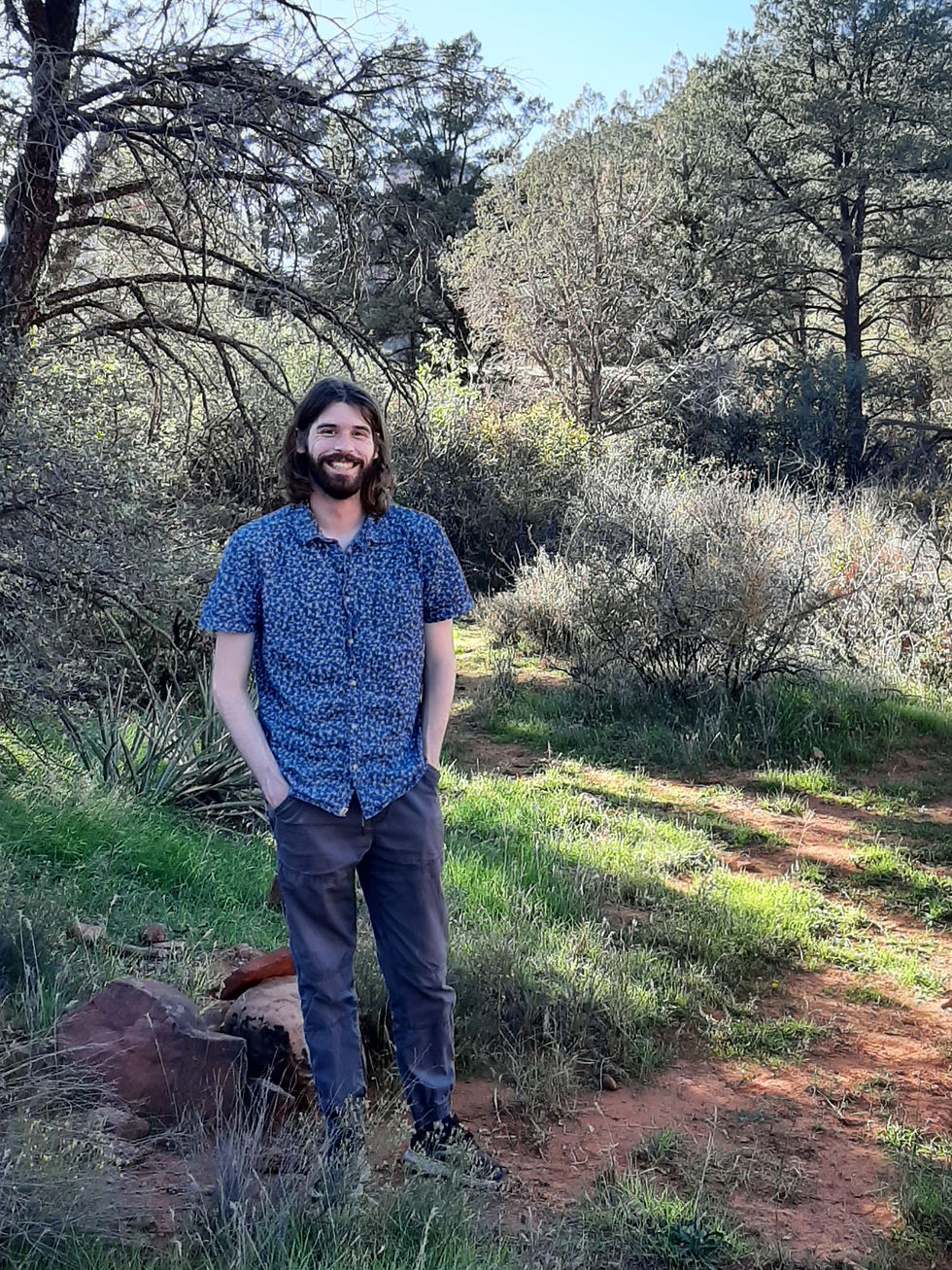 Man smiling in a blue patterned shirt stands in a sunny forest clearing with green grass and tall trees in the background.