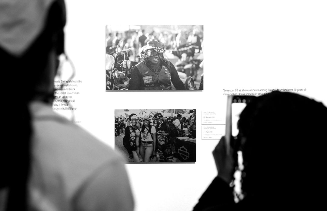 People viewing photographs; two images on wall; one person taking photo.