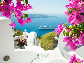 Whitewashed buildings and winding stairways in Santorini frame a breathtaking view of a luxury cruise ship sailing through the deep blue Aegean Sea. Vibrant pink bougainvillea blossoms surround the scene, creating a colorful foreground against the iconic Greek island landscape under clear sunny skies.
