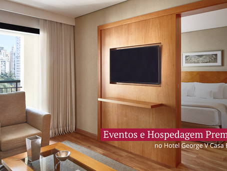SuÃte do Hotel George V Casa Branca com área de estar elegante, poltrona ao lado de uma grande janela com vista para a cidade, televisão em painel de madeira e quarto com cama king-size ao fundo, transmitindo conforto e hospedagem premium.