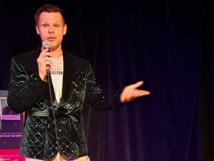 Aaron holds a microphone, performing stand-up. He wear light coloured trousers and a green smoking jacket