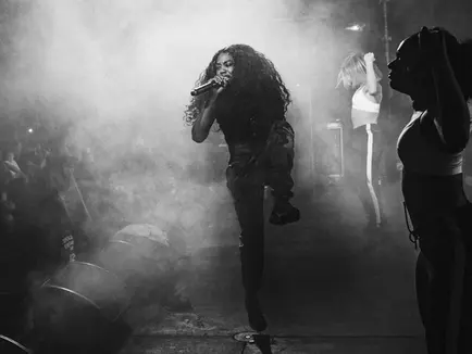 Image shows a black and white photo of Lady Leshurr performing on stage, either side of her are two dancers, the audience can be seen in the background and smoke machines have been used. 