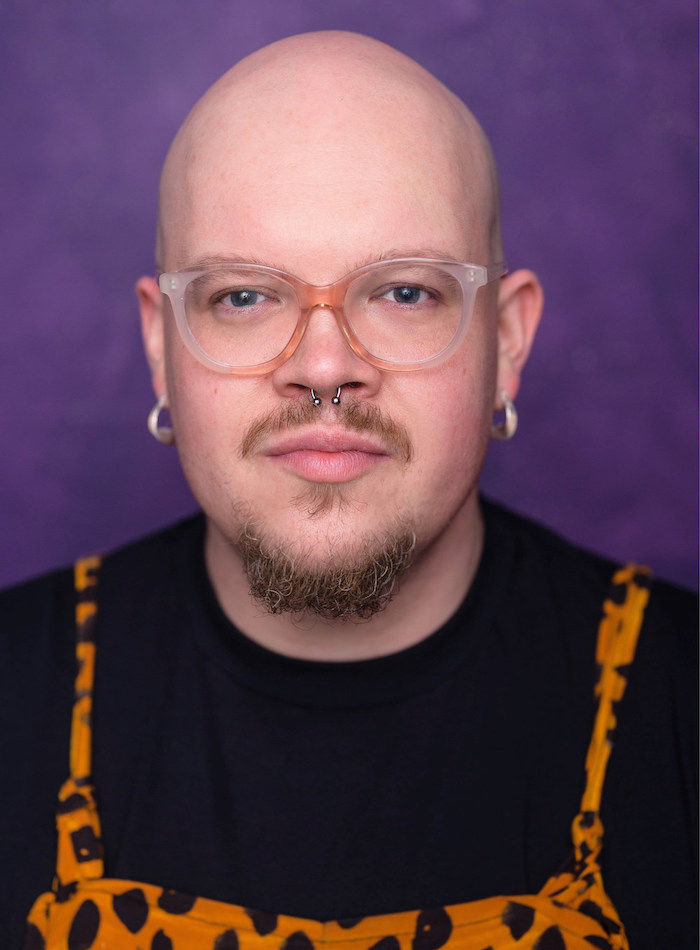 Headshot for Adam Carver. They are a White person wearing glasses, a black top, dungarees and silver earrings. They have a beard, mustache and are bald.