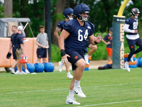 Chicago Bears player No. 64 in navy practice gear runs on a grassy field, with teammates and blocking pads behind him.