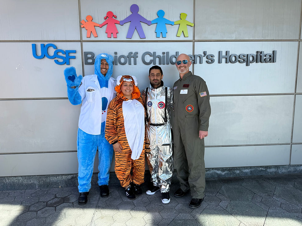 Four people in costumes, including a blue bear and tiger, stand smiling outside UCSF Benioff Children's Hospital. Bright and cheerful mood.
