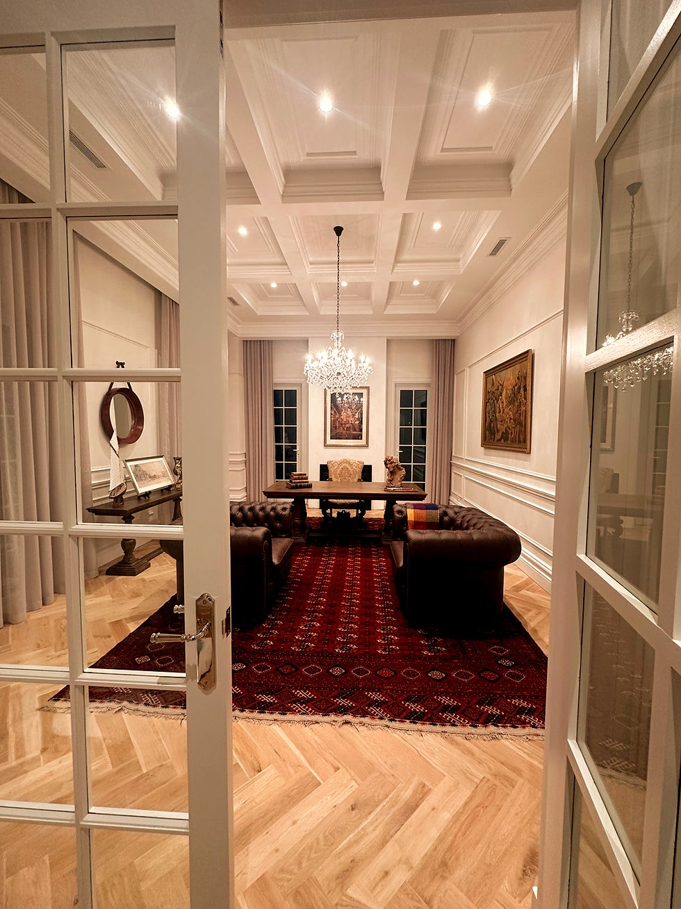 Elegant living room with a chandelier, red patterned rug, and brown sofas. Art on walls, herringbone floor, and open white glass doors.