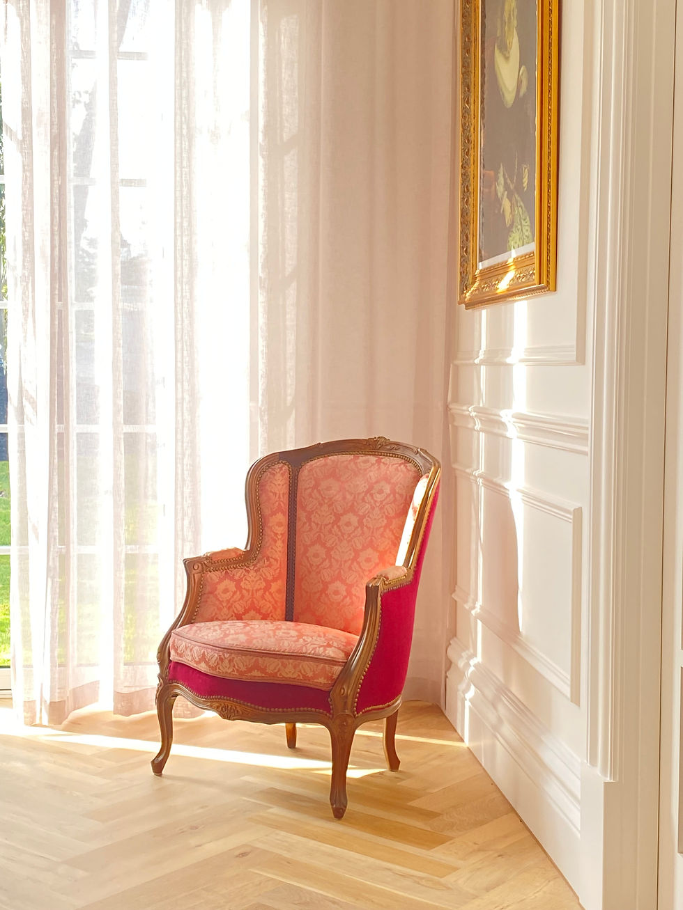Elegant chair with pink floral upholstery in sunlit room, next to sheer curtains and framed artwork on white paneled wall. Warm, serene mood.