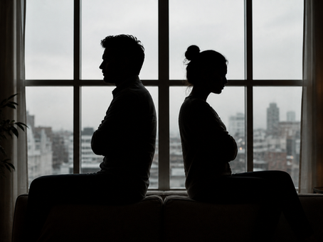 Silhouetted couple sits back-to-back on a couch in front of a window. Mood is tense. Overcast sky outside, warm lamp light inside.