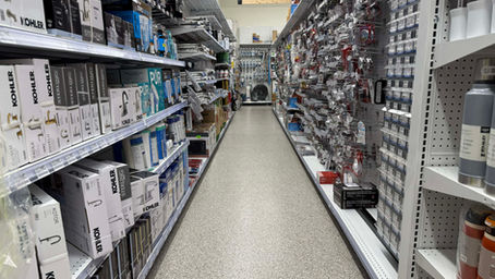 Store aisle with plumbing supplies on shelves. Boxes and fixtures displayed neatly. "PLUMBING" sign visible above. Bright, organized setting.