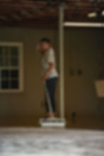 A woman in casual attire spreads epoxy on a floor with a roller. Dimly lit room with a window, pipes, and exposed ceiling. Focused expression.