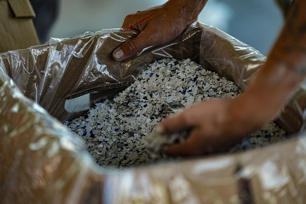 Hands scoop white and blue plastic flakes from a cardboard box lined with plastic, set in an industrial environment.