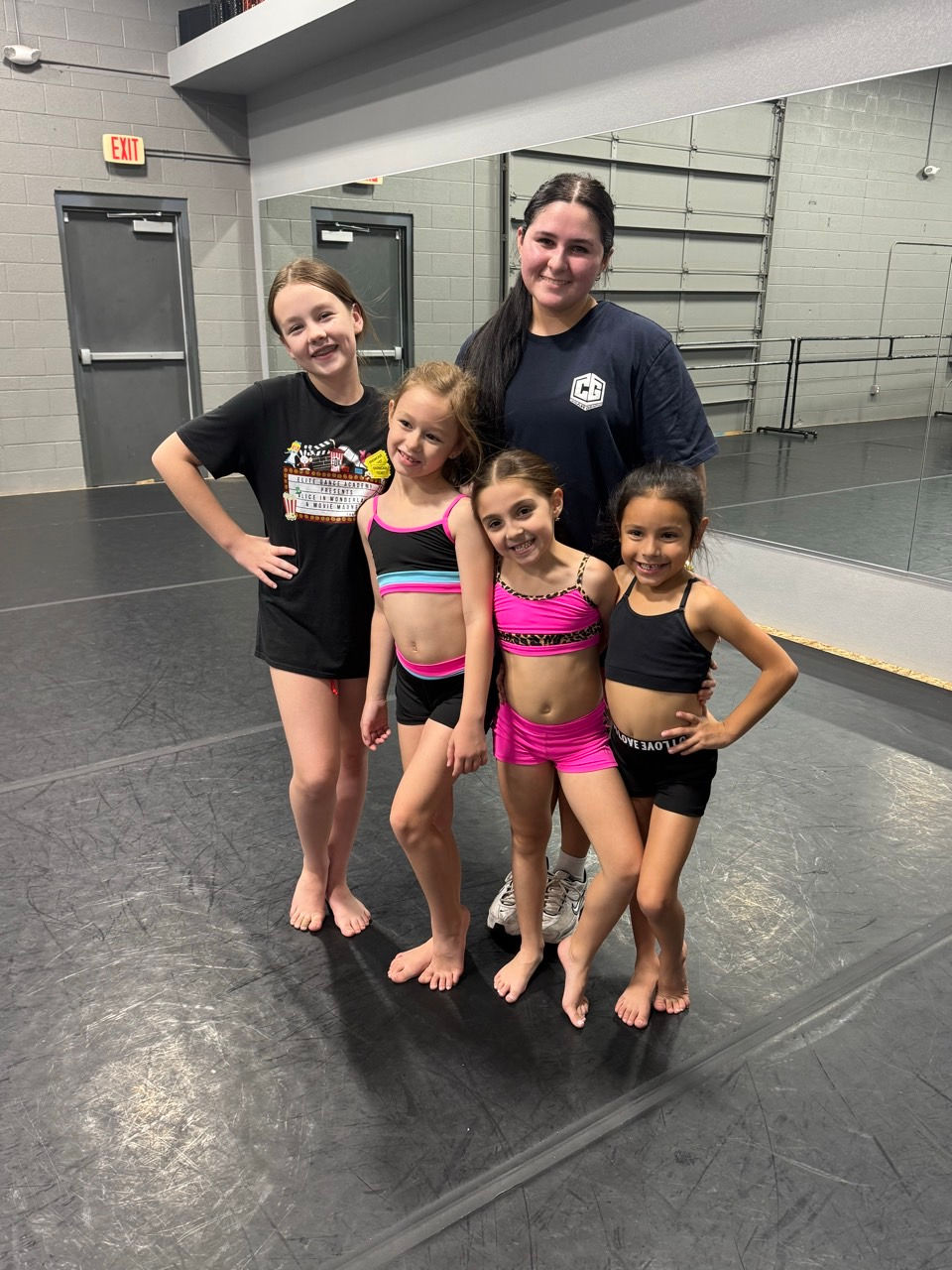 Four smiling girls in dancewear and a woman pose in a dance studio with grey walls and mirrors. A mix of black and pink outfits.