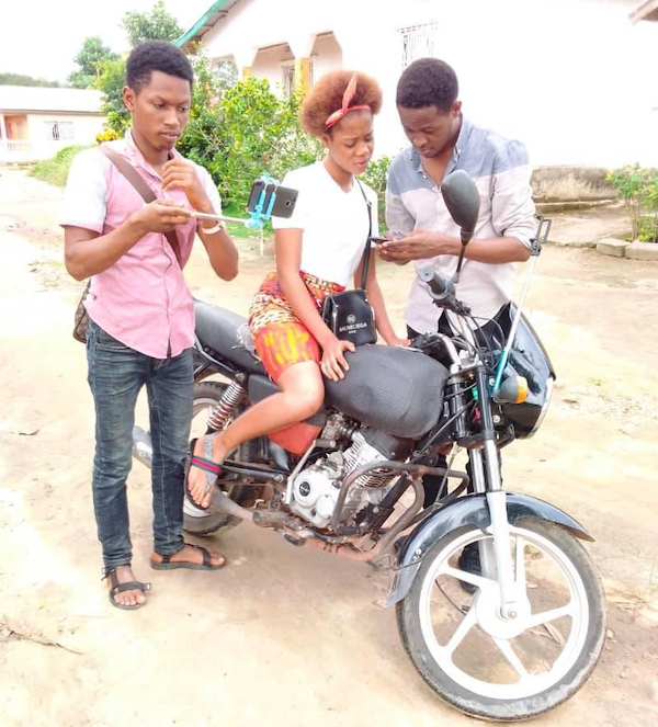 One female on a motorcycle looking at the phone a Male is showing her. While another male appears to take a picture with their mobile phone on a selfie stick.