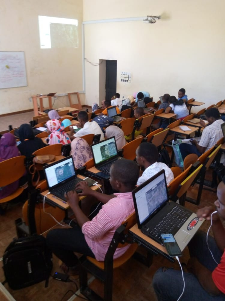 18 students picture in a classroom sitting at their desks, many are on computers with aerial imagery showing.