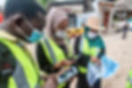 Three individuals (one male two female) wearing safety vests and masks looking at a map in a community street. Two of the individuals looking at the map also have phones.