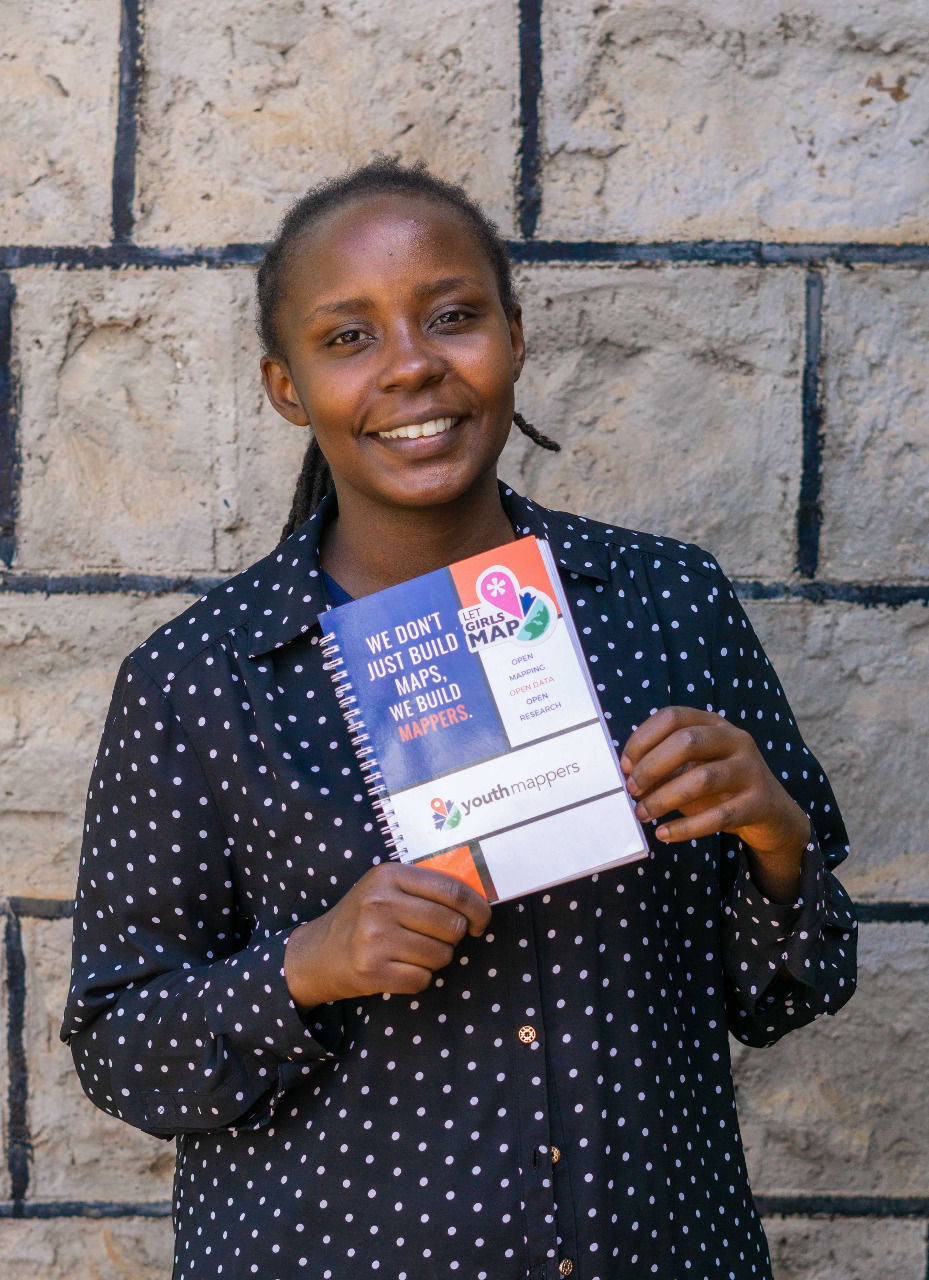 Portrait of Joy, female, in a black and white polka dotted dress shirt holding a YouthMappers journal.