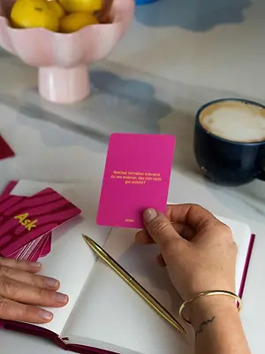 Hand hält eine pinke finimi Fragekarte mit Reflexionsfrage über einem offenen Notizbuch auf einem Schreibtisch.