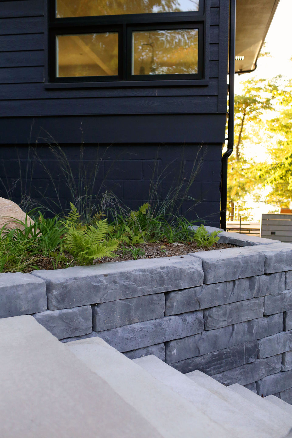 a stone wall with plants growing on it in front of a black house