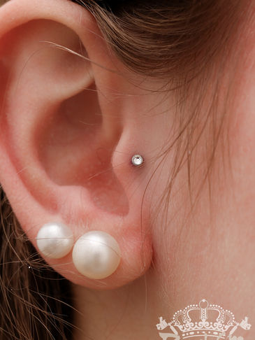 Weibliches Ohr mit Perlen-Ohrstecker und ein Piercing mit Glitzerstein am mittleren Ohrknorpel.