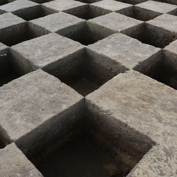 An image of many square test pits dug into the ground with the light greyish-brown soil contrasting with the shadowed depths of the pits to make it look like a chess board. 