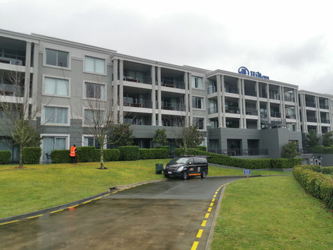 The image shows a man in a fluoro vest cleaning the exterior of the Hilton Hotel, whilst a black work van is parked in the foreground.