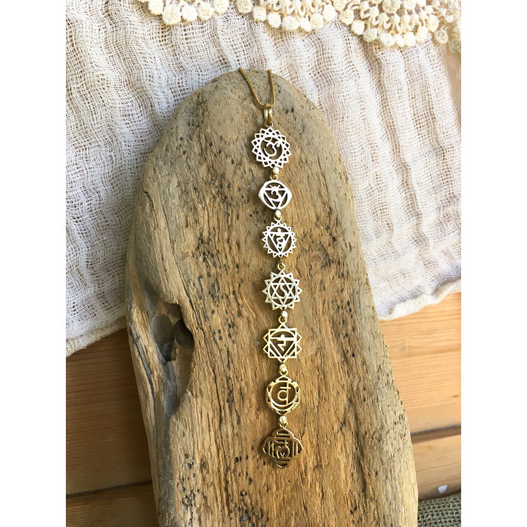 Brass chakra necklace hanging vertically from a piece of driftwood against a wooden background with soft cotton fabric.