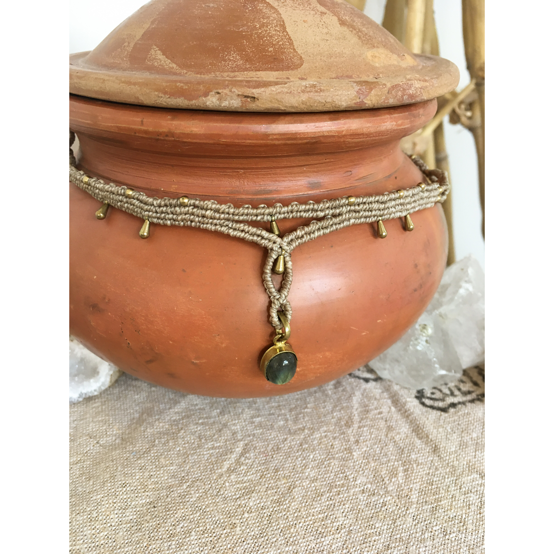 Macramé labradorite necklace displayed on a rounded terracotta pot against a neutral beige fabric background with natural cry