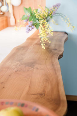 Foto einer Küchentheke aus einem Holzstamm. Die Form ist natürlich gehalten und die Optik des Holzes blieb bestehen. Ein Strauß Flieder steht aus Dekoelement auf der Theke.